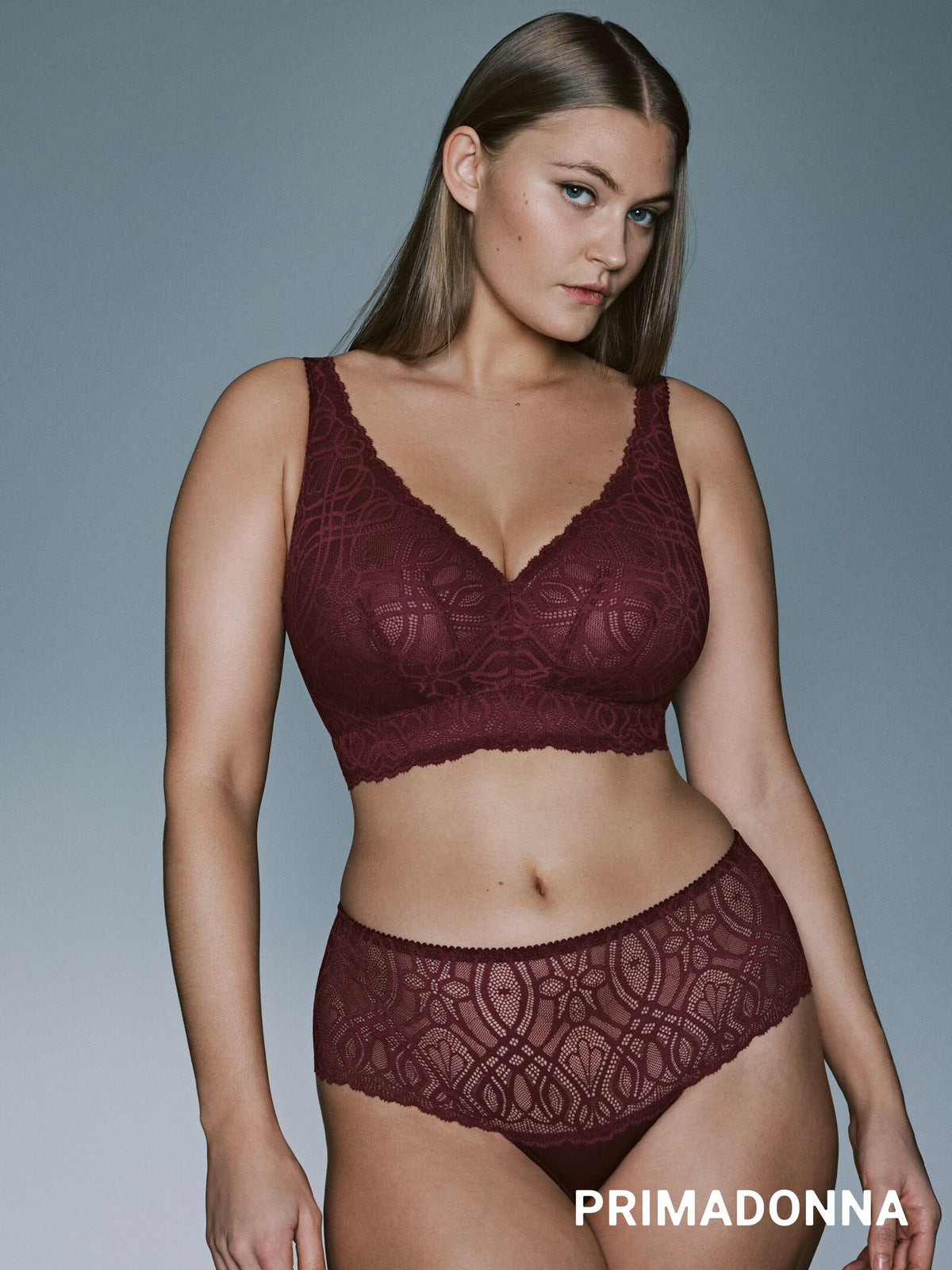 A woman modeling a matching burgundy lace bralette and high-waisted brief set against a solid gray studio background. The brand name PRIMADONNA is printed in white text at the bottom right.