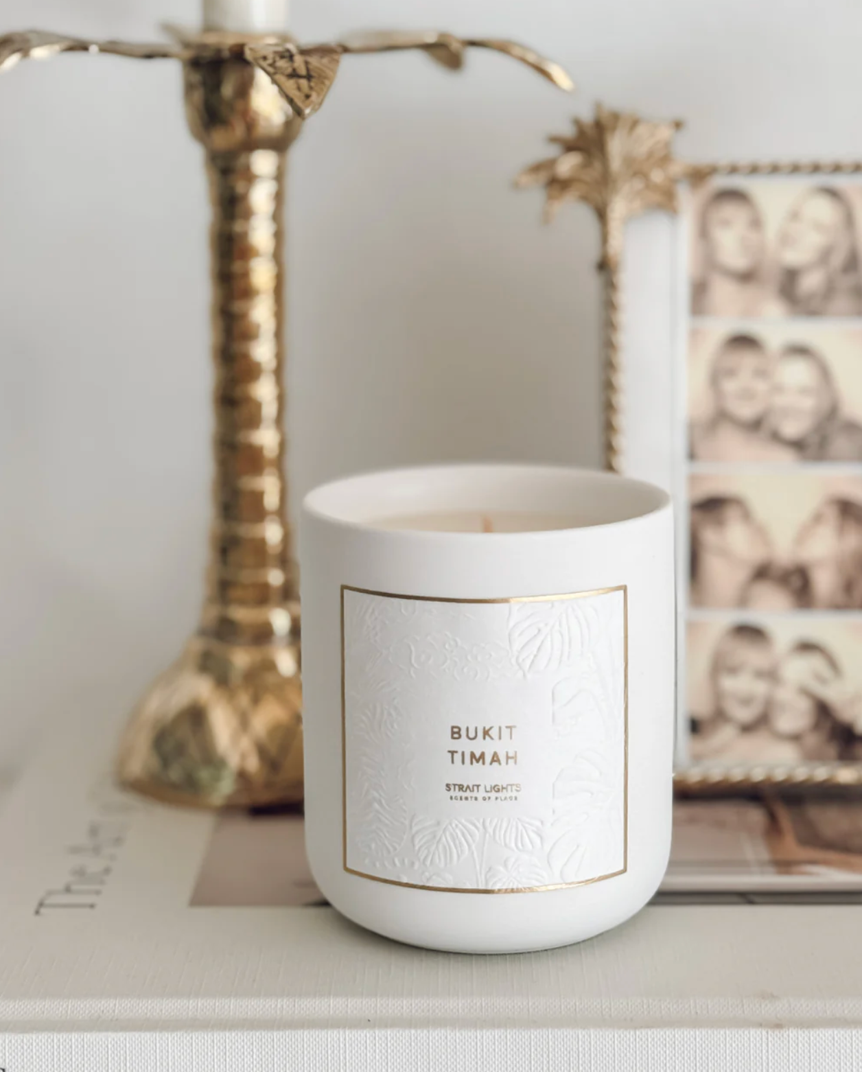 White candle with a label on a surface, with a decorative frame and gold candle holder in the background.