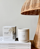 Candle and packaging labeled 'Holland Village' on books with a wicker lamp in the background.